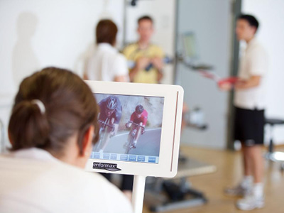 Frau sitzt auf einem Fahrrad-Trainingsgerät und verfolgt das virtuelle Radfahrtraining auf einem Bildschirm im Sportmedizinsaal.