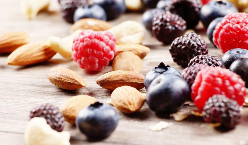 Frische Beeren wie Himbeeren, Blaubeeren und Brombeeren mit Mandeln und anderen Nüssen auf einem Holztisch verstreut.