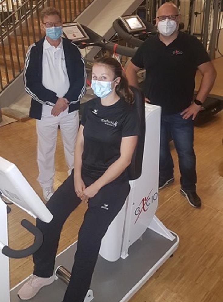 Personen bei einer Demonstration des Pelvictrainer-Geräts, eine Person sitzt auf dem Gerät und wird von zwei weiteren Personen beobachtet, alle tragen Masken.