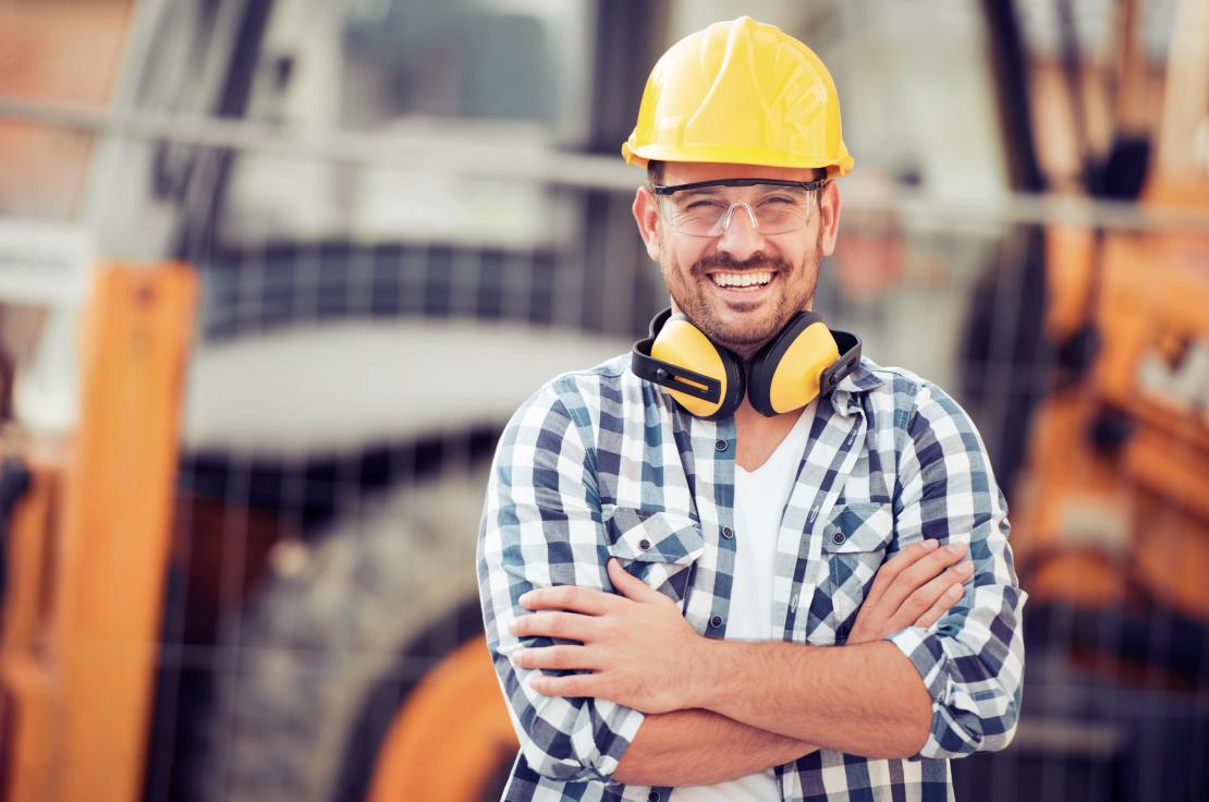 Bauarbeiter in Schutzhelm und kariertem Hemd steht selbstbewusst auf einer Baustelle mit verschränkten Armen.