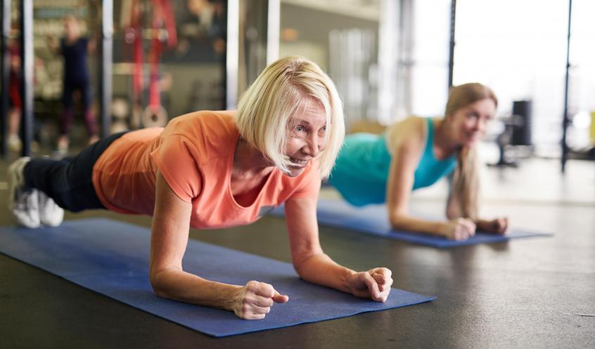 Ältere Frau macht eine Plank-Übung auf einer Matte im Fitnessstudio, begleitet von einer anderen Frau.