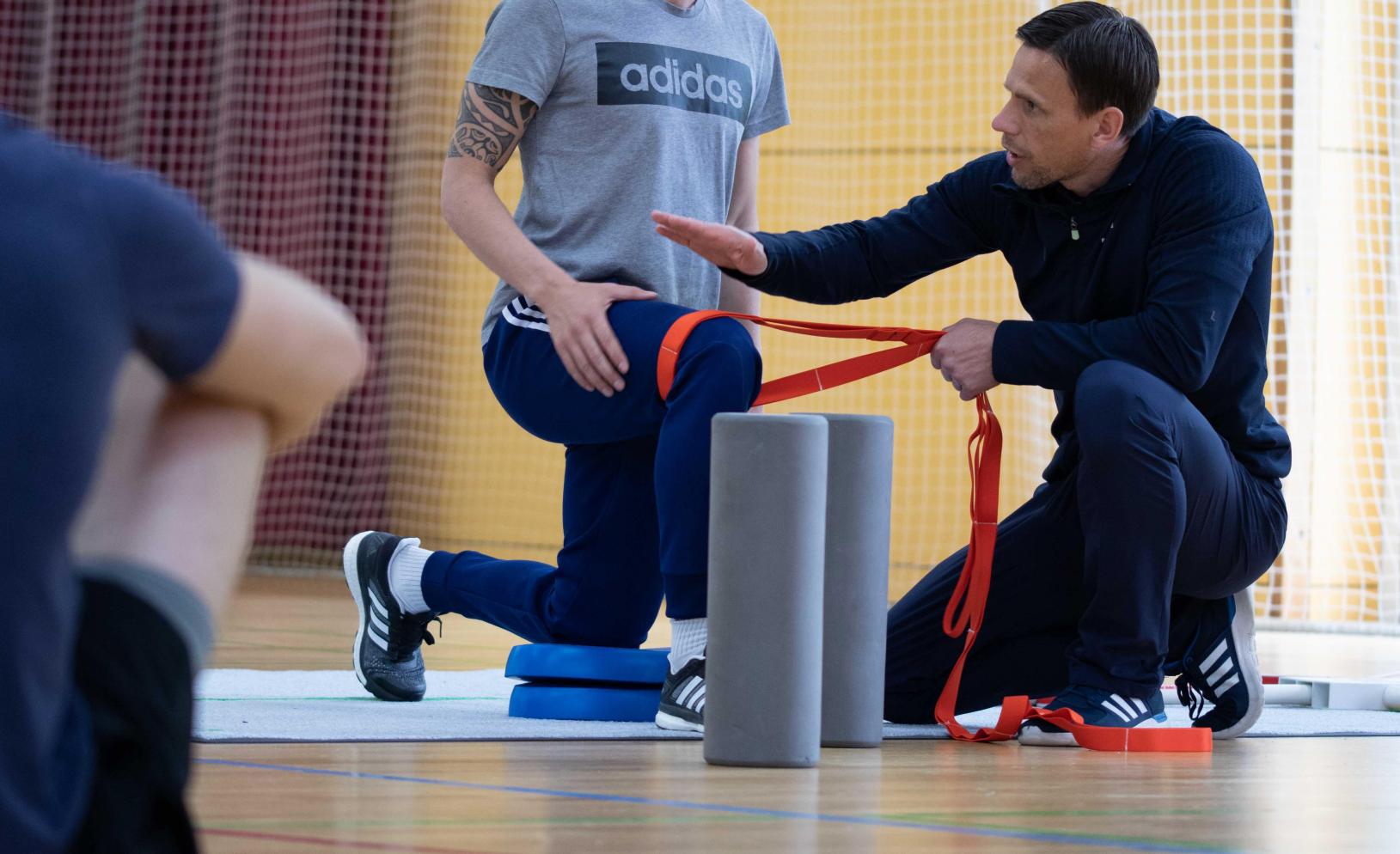 Trainer erklärt einem Sportler eine Übung mit Theraband in der Halle.