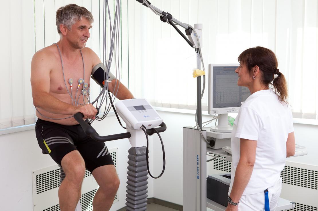 Mann auf Ergometer beim Belastungs-EKG, überwacht von einer Ärztin in einem medizinischen Raum.