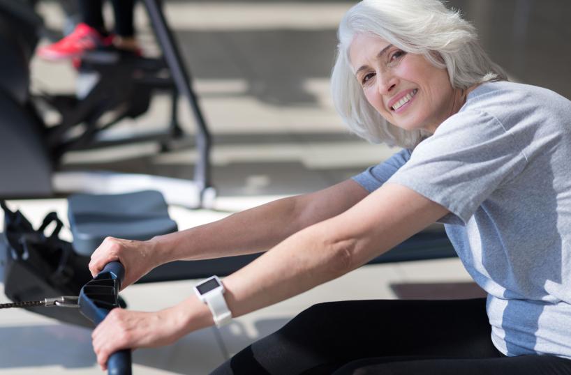 Eine ältere Frau lächelt, während sie an einem Fitnessgerät trainiert, offensichtlich in einer Reha-Einrichtung.