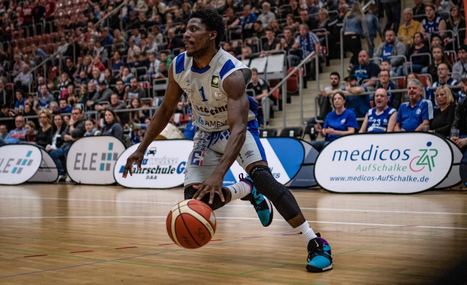 Ein Basketballspieler in Aktion während eines Spiels, im Hintergrund ist das medicos-AufSchalke-Logo sichtbar.